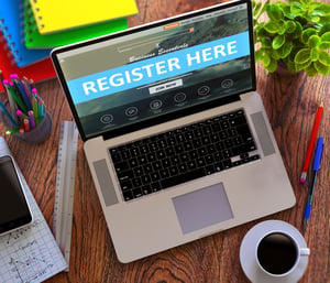 Register Here Concept. Modern Laptop and Different Office Supply on Wooden Desktop background.