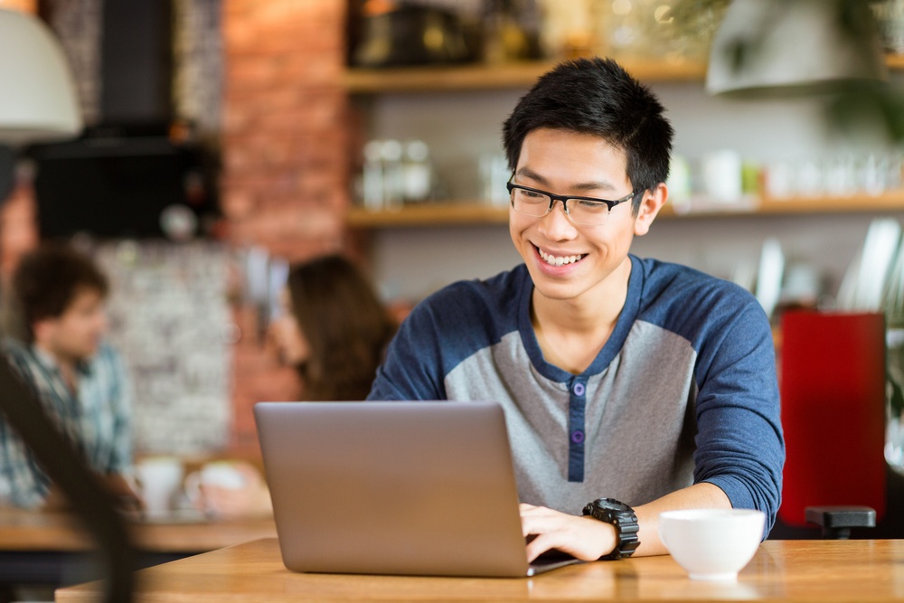 Happy cheerful young asian male in glasses smiling and using laptop in cafe-1.jpeg